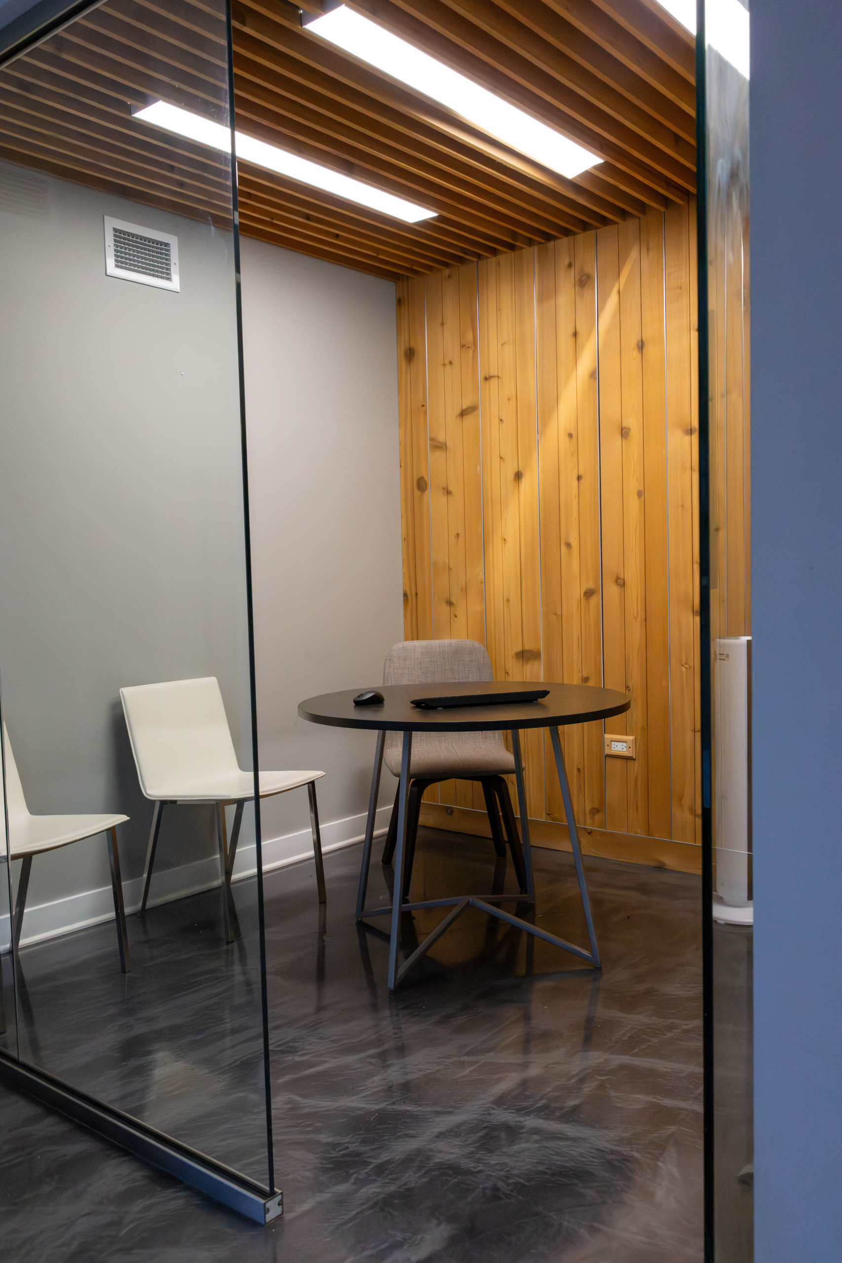 Small glass-walled meeting room with a round table, three chairs, and wood-paneled accent wall